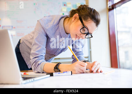 Smiling pretty young woman drawing blueprint on the table near the window Stock Photo