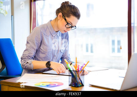 Concentrated pretty young woman fashion designer sitting and drawing sketches in office Stock Photo
