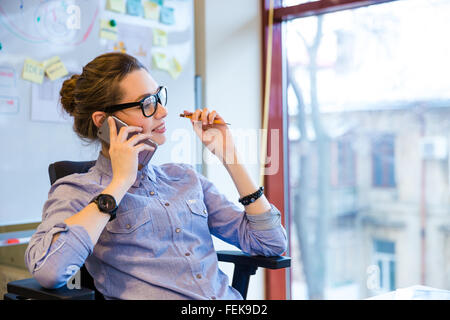 Happy beautiful young business woman in glasses sitting and talking on cell phone in office