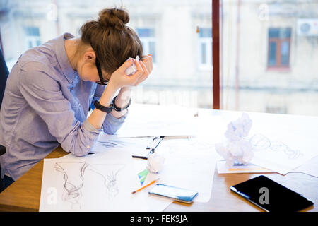 Sad despaired young woman fashion designer sitting in office with sketches on table
