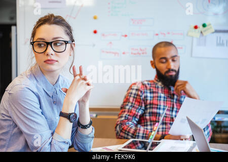 Pensive young woman in glasses sitting in conference room and thinking while her male colleague reading Stock Photo