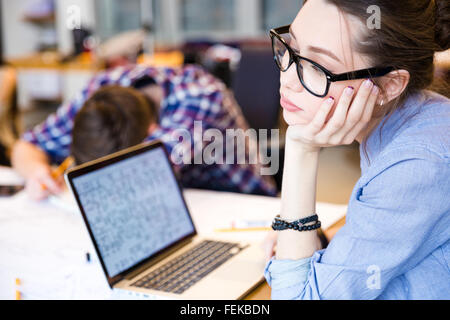 Tired young woman in glasses with laptop sitting in meeting room while her colleague sleeping on the table Stock Photo