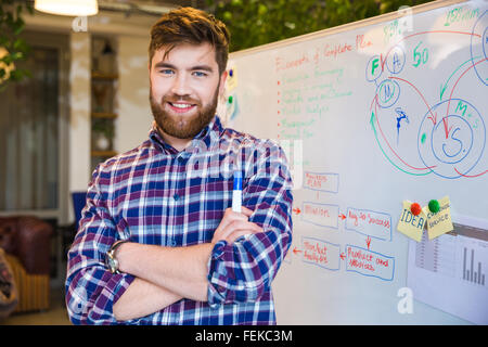 Happy young businessman standing in office with whiteboard on background and looking at camera Stock Photo