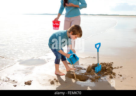 Little boy playing on the beach with a bucket and spade with his mother. Stock Photo
