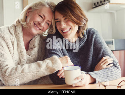Laughing mother and daughter in sweaters hugging and drinking coffee Stock Photo