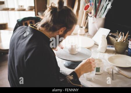 Rear view of mid adult man in workshop applying ceramic glaze to clay pot Stock Photo