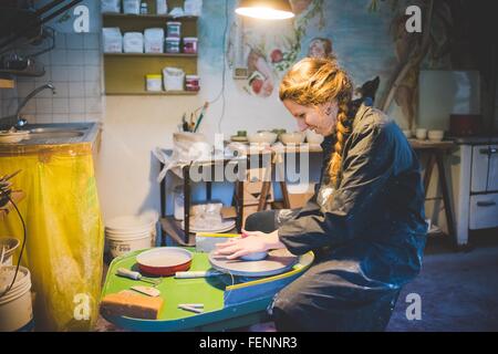 Side view of young woman sitting in workshop using potters wheel, looking down smiling Stock Photo