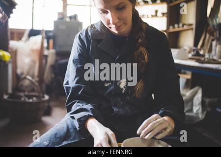 Front view of young woman sitting at pottery wheel looking down making clay pot Stock Photo