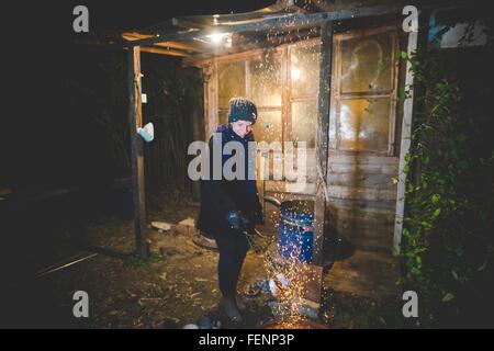 Young woman by shed waving tongs over fire causing sparks, looking down smiling Stock Photo