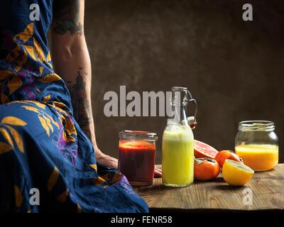 Cropped view of  woman wearing dress sitting on table with raw juices in glass bottle and jars Stock Photo