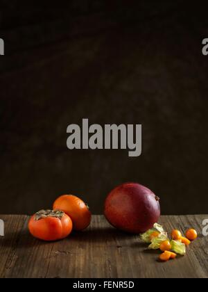 Whole mango and persimmons on wood table against dark background Stock Photo