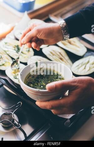 Mans hands drizzling herb oil on aubergine halves in kitchen Stock Photo