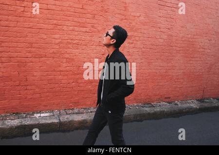 Side View Of Fashionable Young Man Walking On Street Against Red Brick Wall