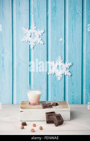 Chocolate cubes, hazelnuts, glass of milk, paper snowflakes on blue wall Stock Photo