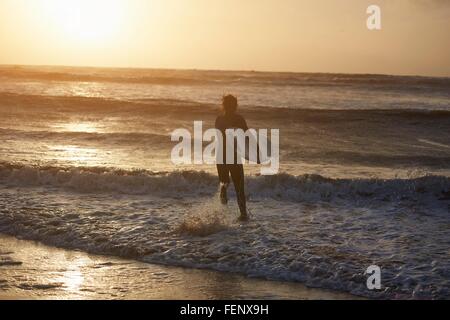 Young male surfer carrying surfboard running into sea, Devon, England, UK Stock Photo