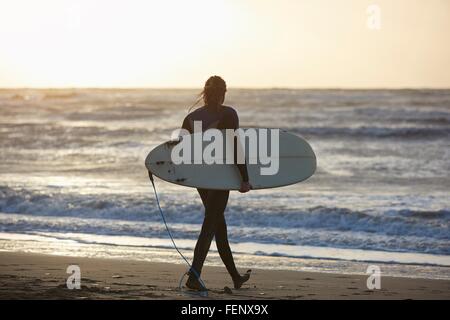 Young male surfer walking on beach carrying surfboard, Devon, England, UK Stock Photo