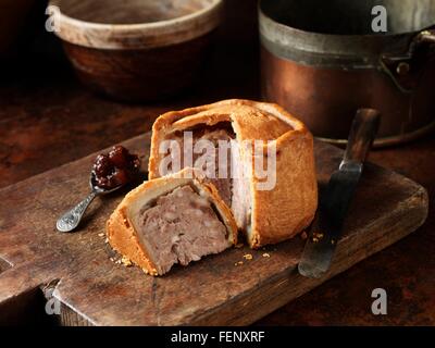 Sliced Melton Mowbray pork pie with spoonful of pickle on wooden cutting board Stock Photo