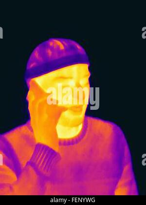Thermal image of young man wearing face mask and hat using smartphone Stock Photo