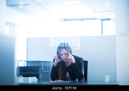 Young woman sitting at desk in office, head in hands looking down, stressed Stock Photo