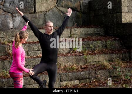 Mid adult man on stone steps arms raised standing on one leg, young woman helping Stock Photo