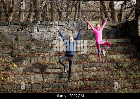 Full length front view of couple on stone steps, arms raised standing on one leg Stock Photo
