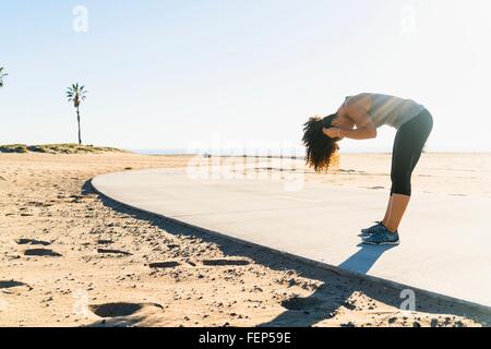 Mid adult woman standing on pathway at beach, bending over, fixing hair Stock Photo