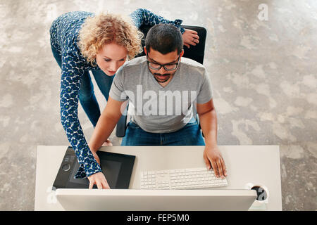 Top view shot of young man sitting at his desk working on pc and digital graphics tablet with woman pointing at monitor. Male gr Stock Photo