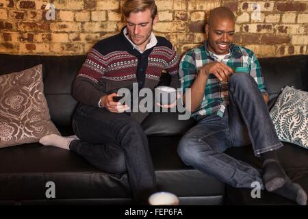 Male couple sitting on sofa, looking at smartphones Stock Photo