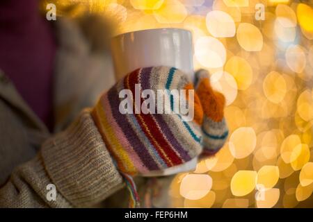 Close up of woman hands wearing mittens holding hot drink in front of xmas lights Stock Photo