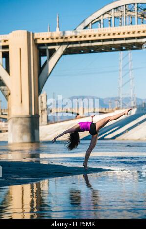 Side view of dancer, leg raised, bending over backwards in front of bridge, Los Angeles, California, USA Stock Photo