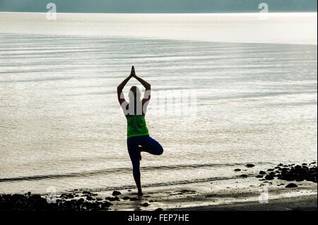 Silhouetted rear view of young woman practicing yoga standing tree pose on beach Stock Photo