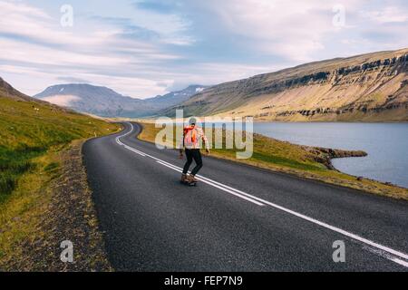 Rear view of mid adult man skateboarding on curving open road by lake, Iceland Stock Photo