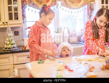 Boy in kitchen wearing santa hat making cookies with sisters peeking over kitchen counter open mouthed Stock Photo