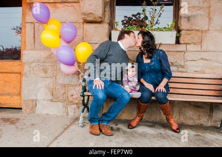 Mature couple sitting on bench, kissing, young daughter sitting between them Stock Photo