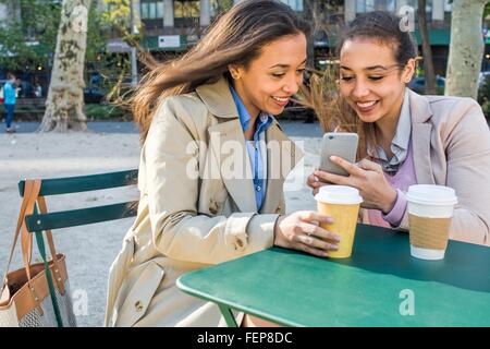 Young adult female twins with takeaway coffee reading smartphone texts in city park Stock Photo