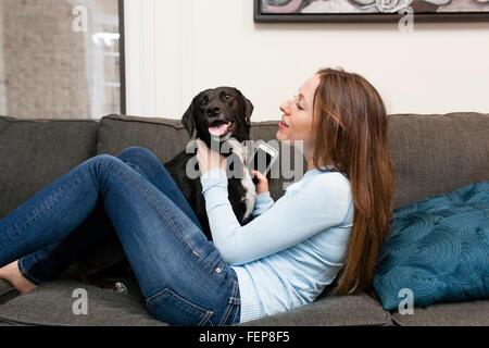 Side view of young woman on sofa holding smartphone playing with dog Stock Photo