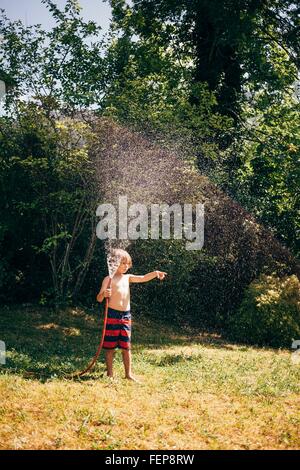 Full length view of boy in garden holding hosepipe spraying water, looking away pointing, Bludenz, Vorarlberg, Austria