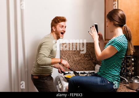 Side view of young woman in kitchen using smartphone to take photograph of young man cooking, open mouthed smiling Stock Photo