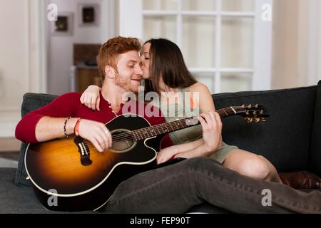 Young woman on sofa kissing young man playing guitar on cheek Stock Photo