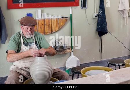 Potter wearing flat cap sitting at pottery wheel shaping clay vase Stock Photo