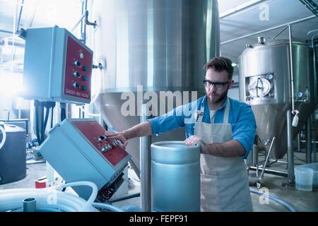 Young man in brewery by conical fermentation tank holding canister, pressing button on control panel Stock Photo