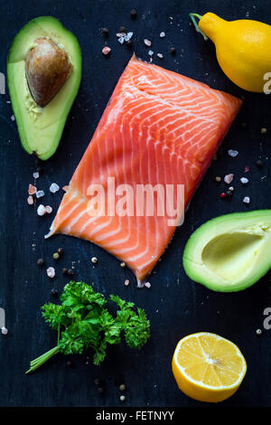 Fresh salmon fillet, lemons, avocados, parsley and spices on black cutting board on textured wooden background. Top view Stock Photo