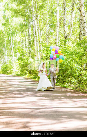 Beautiful bride holding bunch of balloons in the park. Couple of bride and groom with balloons. Newlyweds with balloons outdoors Stock Photo