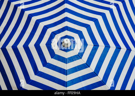 A blue and white striped umbrella set against a bright outdoor backdrop. The geometric pattern of alternating stripes creates a vibrant visual contrast. Umbrellas like this are often used for sun protection and as a decorative element in beach or outdoor settings, offering shade and style. Stock Photo