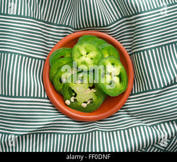 Top view of a small bowl of sliced jalapeño peppers on a green and white wrinkled tablecloth illuminated with natural light. Stock Photo