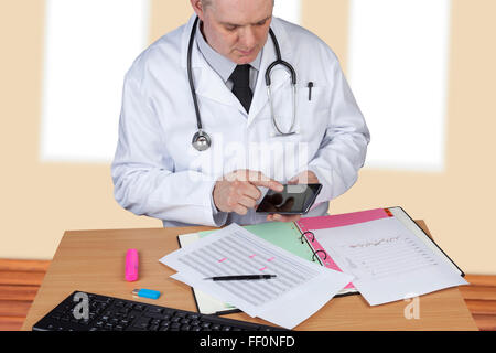 Doctor with stethoscope around his neck using his mobile phone while at his desk checking reports Stock Photo