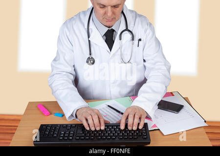 Doctor with stethoscope around his neck typing up a report on his computer at his desk Stock Photo