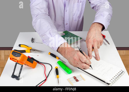 Electronic test engineer writing his circuit testing results in a note pad on his white topped work bench Stock Photo
