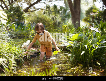 A boy wades knee deep in a small pond in a lush green setting. Stock Photo