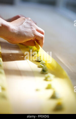 A close-up on hands folding homemade pasta dough over small dots of filling on a wooden counter top. Stock Photo